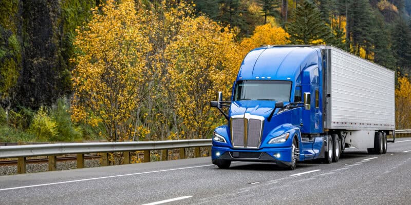 blue freight truck driving in fall foliage on highway